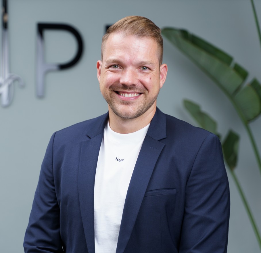 Ein Mann in blauem Anzug und Krawatte steht in einem Haus und lächelt in die Kamera, während Fenster und Licht im Hintergrund verschwimmen.